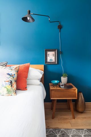 Blue bedroom wall with a wood bed, white duvet set and orange cushions with black wired lampshade