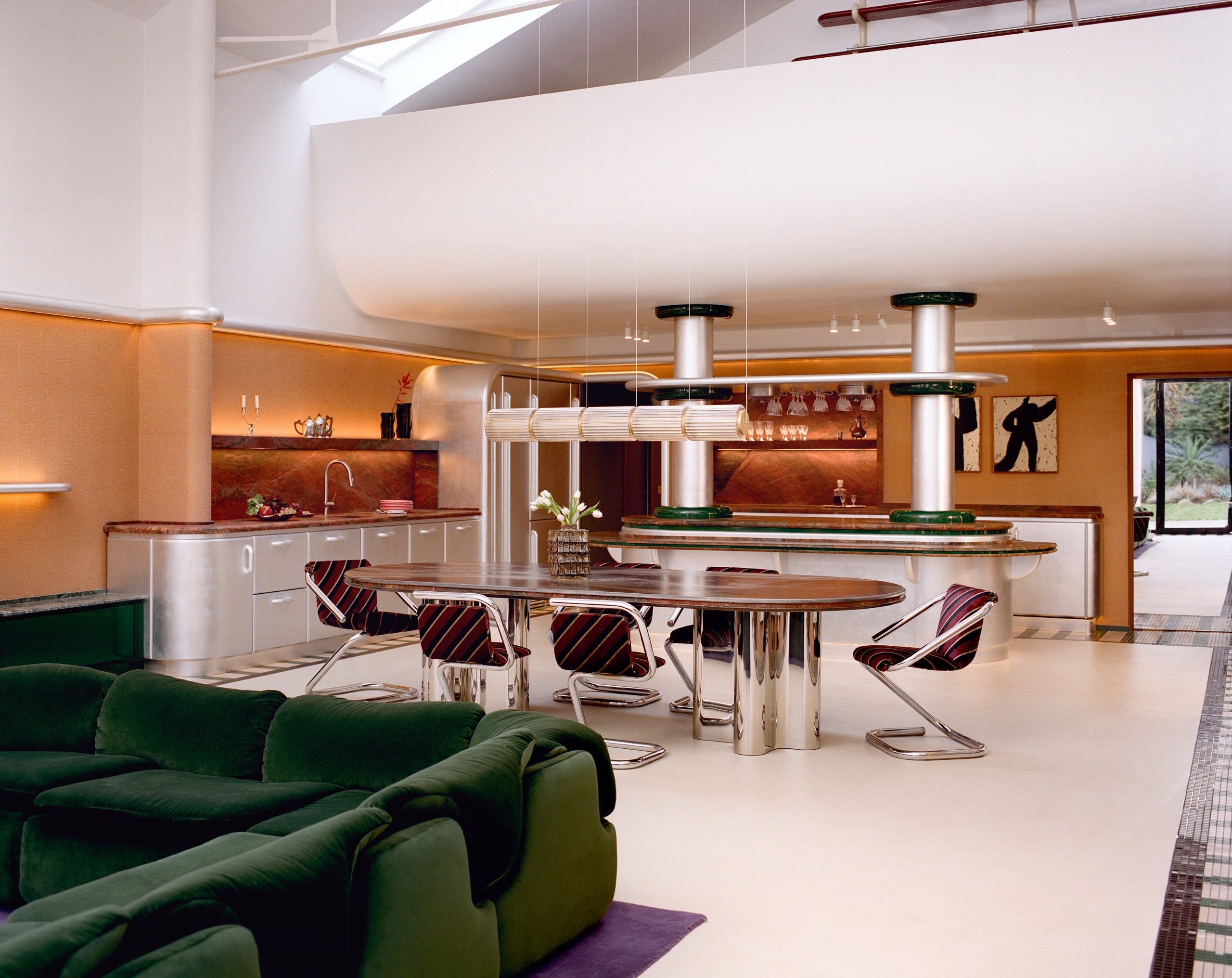 An open living, dining, and kitchen space in a Space Age-inspired home. There is orange wallpaper, a white ceiling, mosaic tiles floors, a dining table with purple chairs and a green sofa