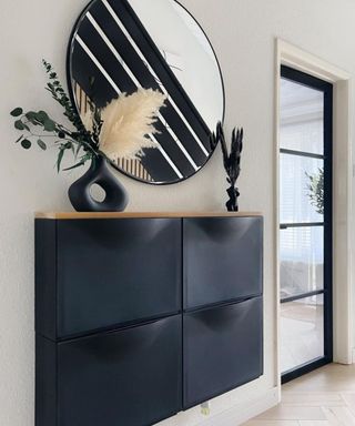 A black IKEA TRONES shoe storage cabinet mounted to wall and decorated with matt black vase. A black framed mirror is positioned above