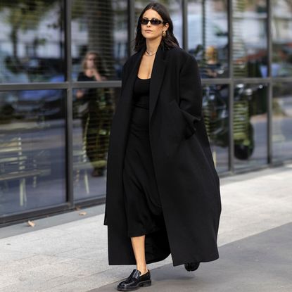 fashion week attendee wearing long black coat, sunglasses, black dress, and loafers