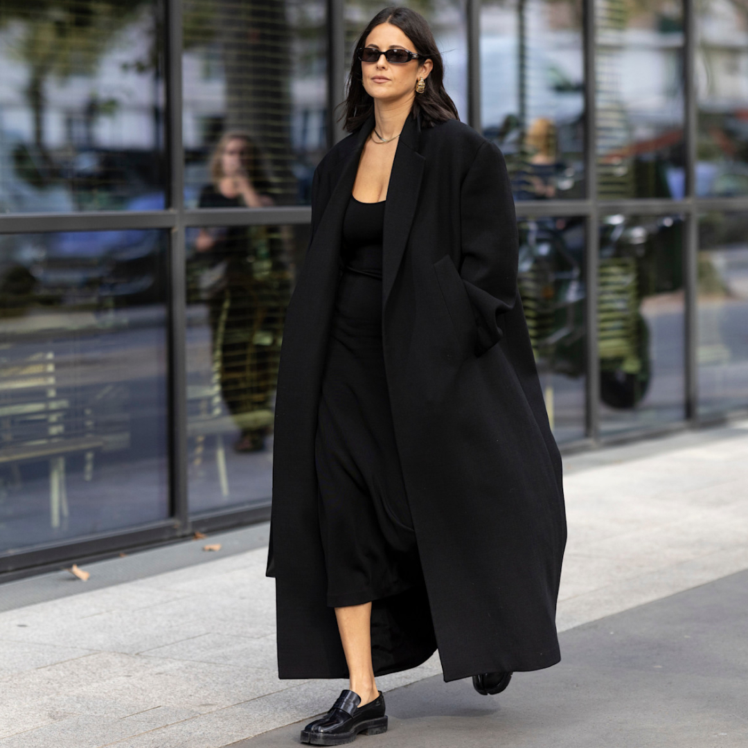 fashion week attendee wearing long black coat, sunglasses, black dress, and loafers