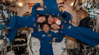four astronauts with their heads together, floating in a circle on the space station