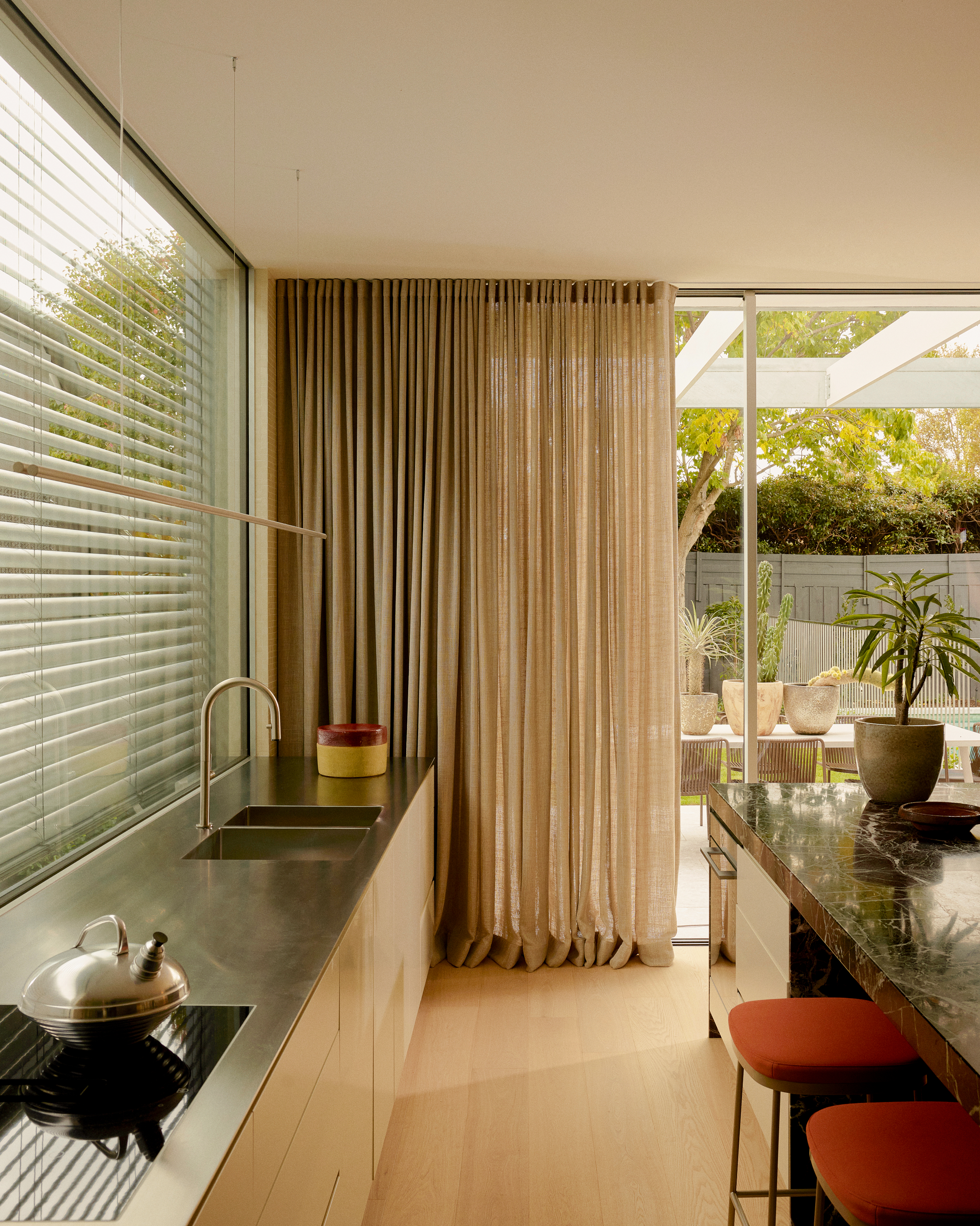 Kitchen with sleek white cabinets, stainless steel worktops, and sheer curtains covering sliding doors out to garden