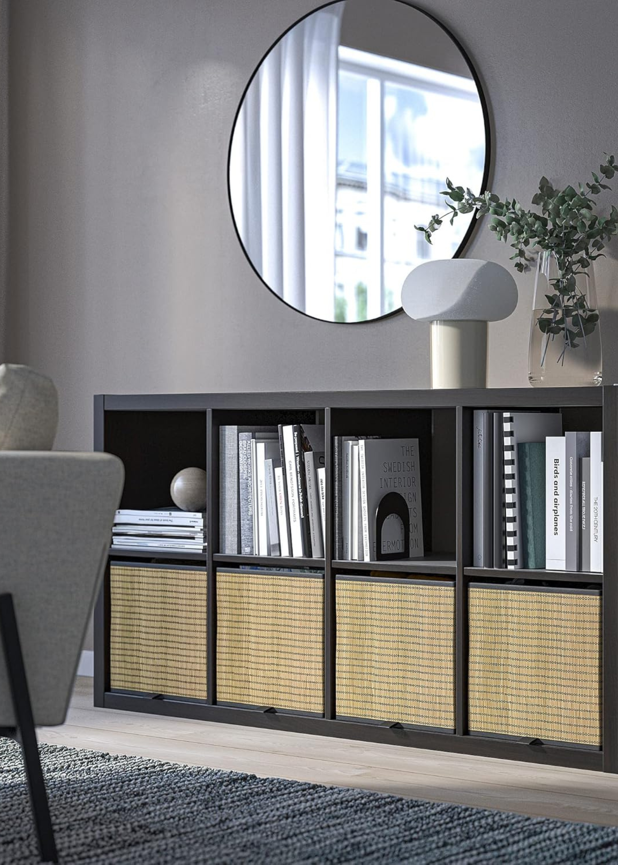ikea kallax unit in dark brown with pullout drawer boxes in the bottom row, books scattered in the top row and a light and foliage on top with a circular mirror on the wall above