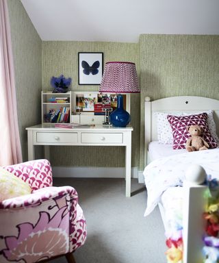 Pale green patterned wallpaper, white desk and bed, and pink printed armchair illustrating girls' bedroom ideas.