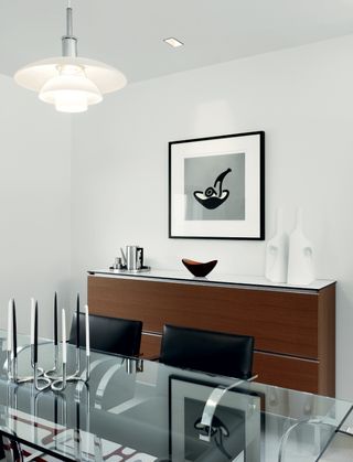 Interior view of Gerry McGovern’s dining room featuring light coloured walls, a pendant light, wall art, a wooden unit with a silver coffee set, black bowl and white sculptures on top, a glass dining table with a silver candlestick holder and black and white candles and black chairs
