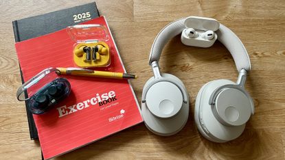 The Cambridge Audio Melomania P100, Sony WF-C710N, AirPods Pro 2 and Nothing Ear (a), which all feature in this student headphones guide, on a wooden table next to a stack of exercise books, a diary and a pen