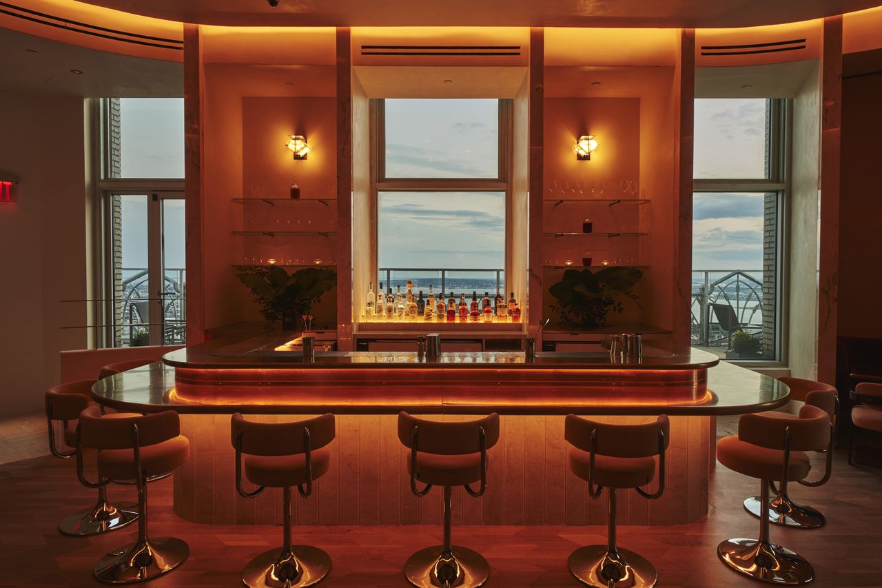 A dimly lit, panoramic bar decorated with 1980s-style chrome and orange velvet chairs, golden-lit sconces, and sleek, back-lit surfaces overlooks a city at dusk.