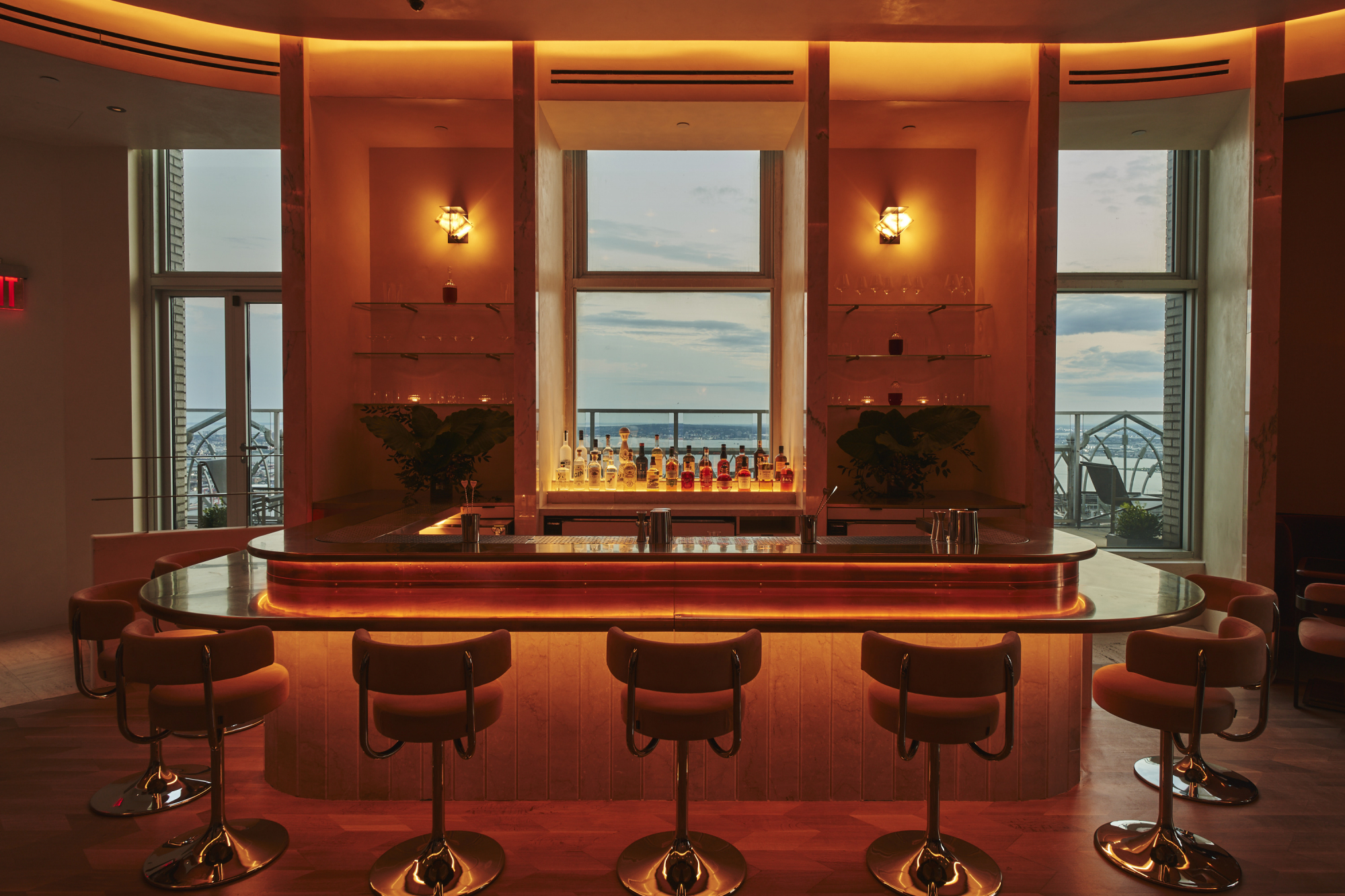 A dimly lit, panoramic bar decorated with 1980s-style chrome and orange velvet chairs, golden-lit sconces, and sleek, back-lit surfaces overlooks a city at dusk.
