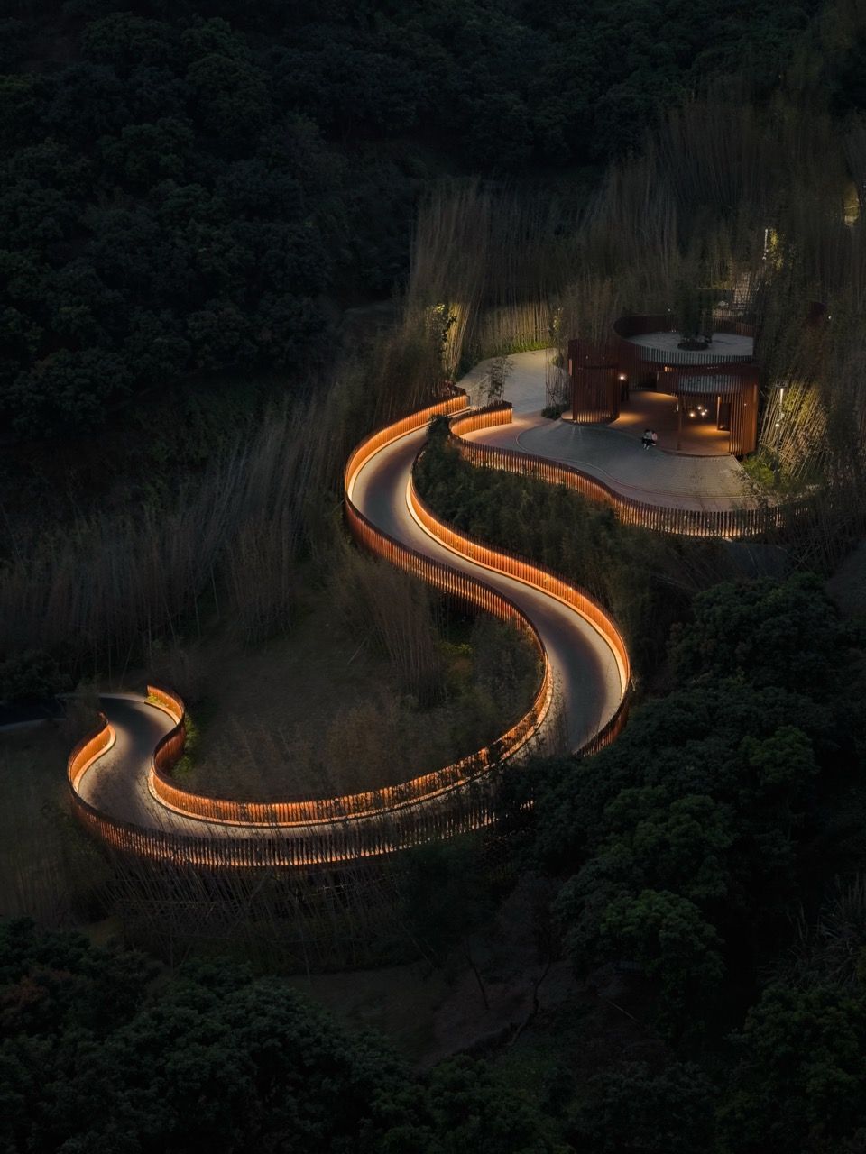 Photograph of a winding driveway leading away from a house, softly lit at night, shot from above.