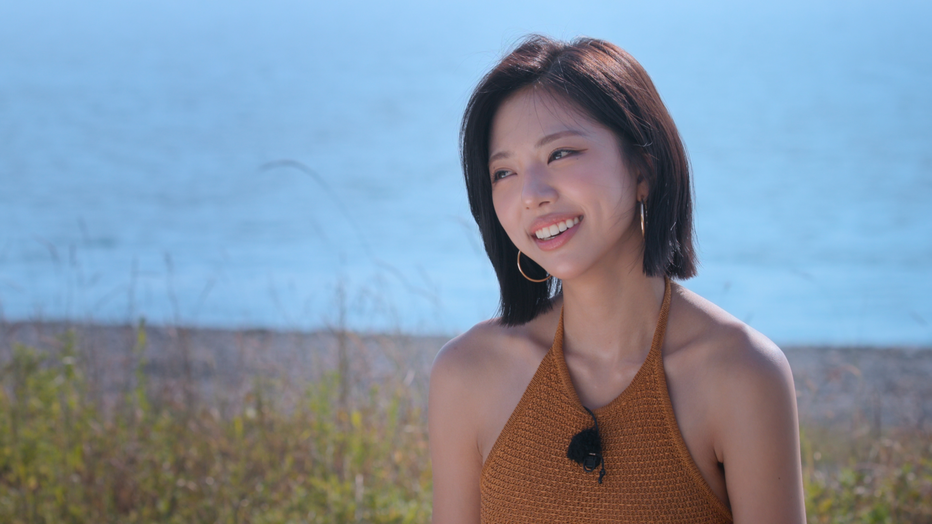 A woman wearing a brown halter top and lav mic (model Kim Go-eun) smiles as she sits in front of a small shoreline, in 'Single's Inferno' season 5.