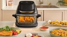 The Cuisinart glass air fryer on a white countertop surrounded by cooked food. 