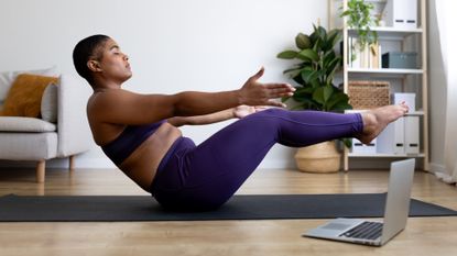 woman performing boat pose in yoga on a grey mat in a living room setting with a laptop to the side of her. 