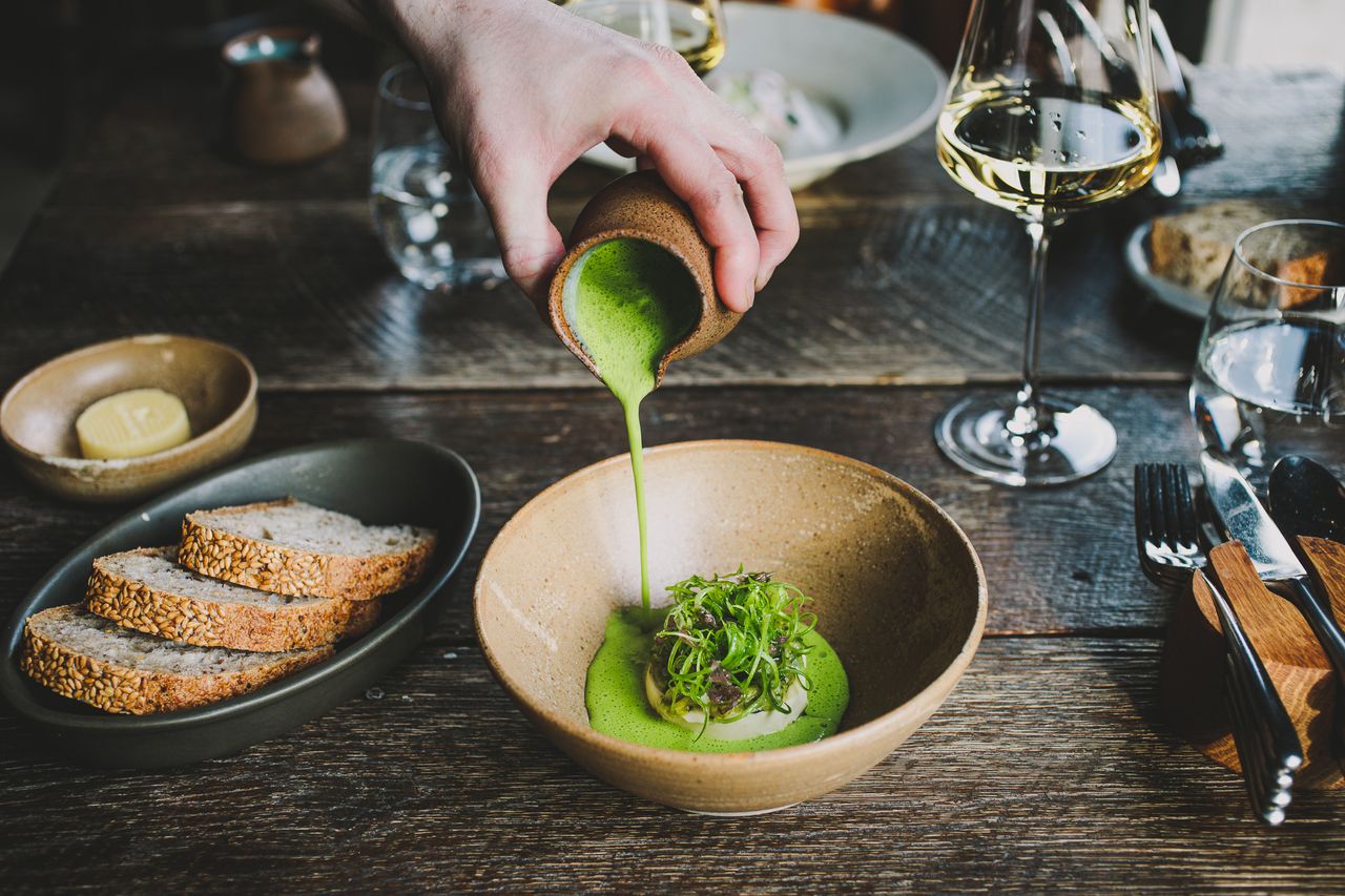 At a sophisticated restaurant, a person completes a vegetable-based plate placed in a crafty ceramic bowl with a green, foamy cream. All around are plates with bread, butter, a wooden cutlery mat, and wine glasses.