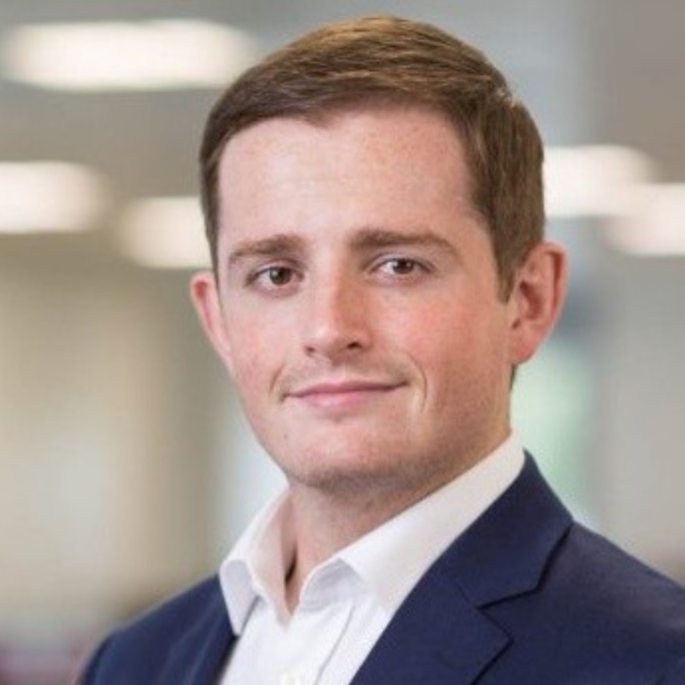 Man with short brown hair wearing a navy blazer and white shirt, headshot with a blurred office background.