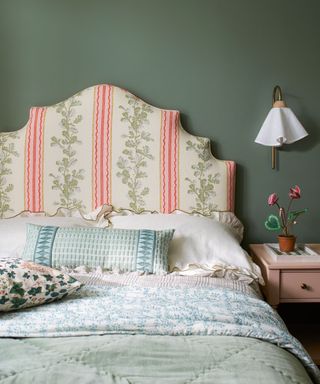 Green painted walls in bedroom, large upholstered headboard with floral fabric, blue, green and white patterned bedding, wooden beside table with wall lamp with fabric shade