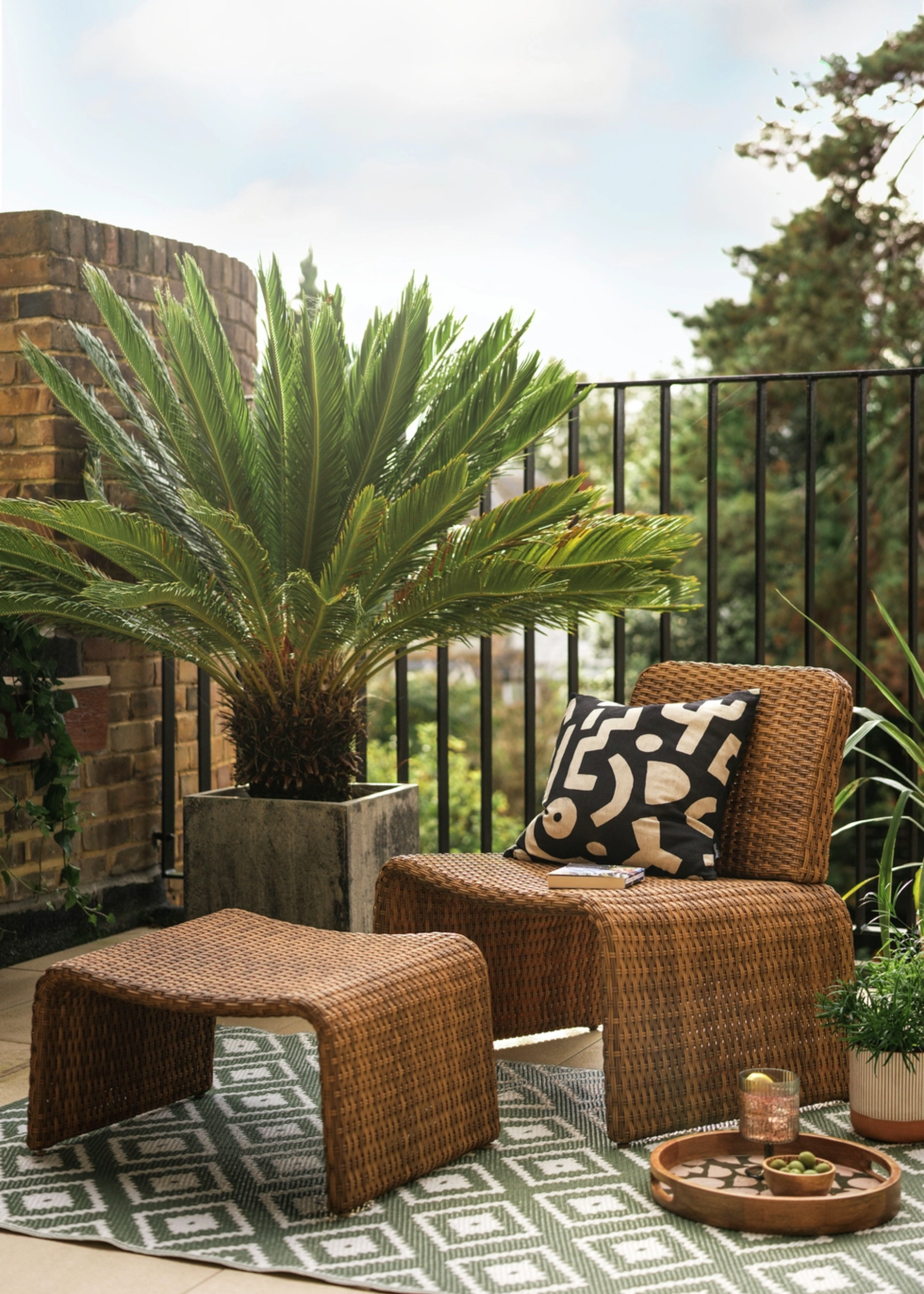 A balcony with a potted dwarf tree, a small rug and a set of woven furniture