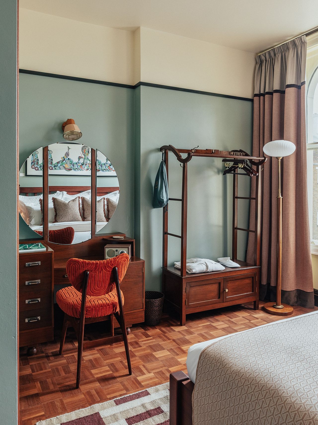 A layered, transitional hotel suite featuring a series of figurative, colorful artworks on its walls, patterned furniture, including rugs, armchairs, and sofas, split-color pillars, and a buttery, tile blue, and green palette.