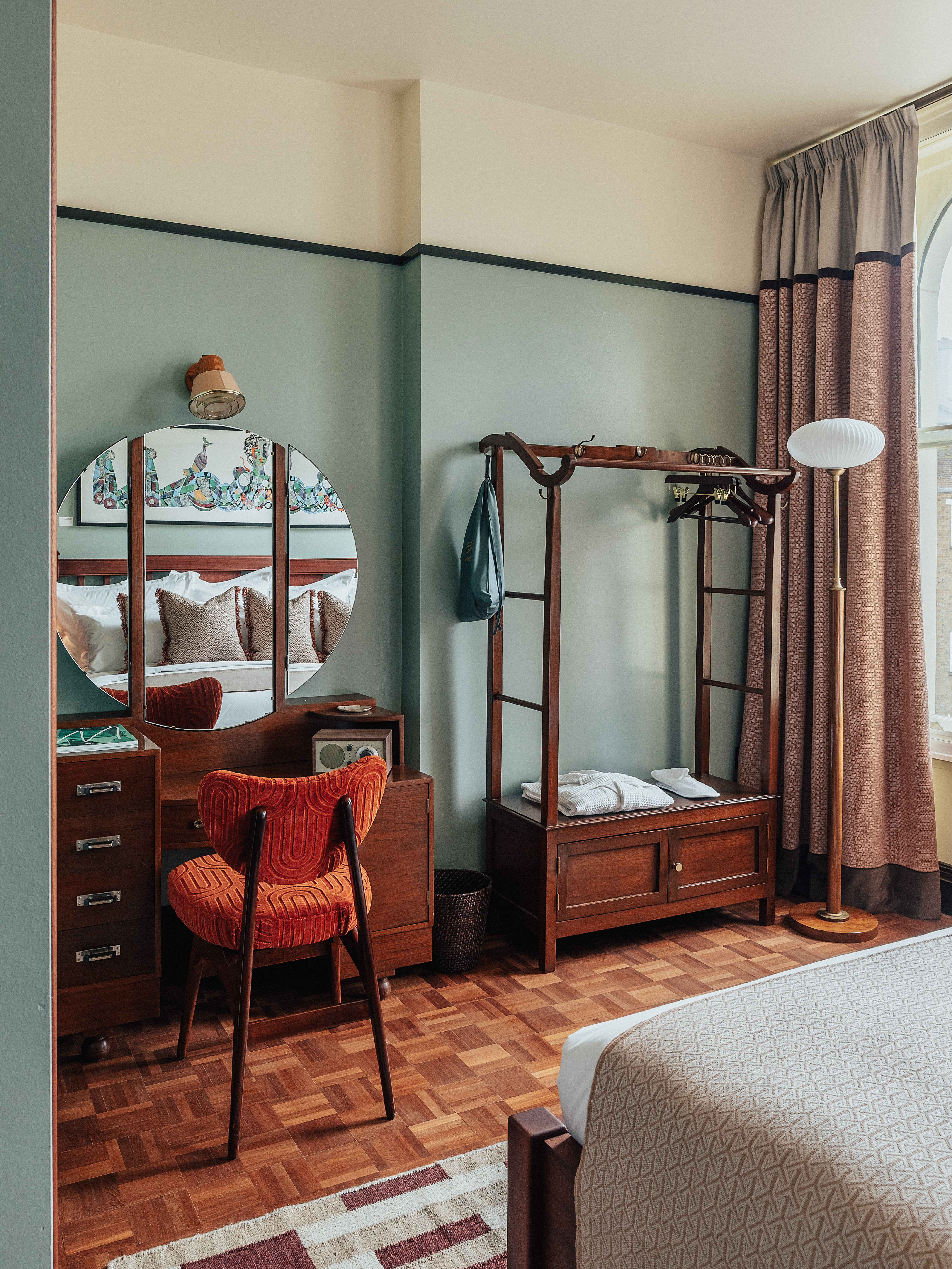 A layered, transitional hotel suite featuring a series of figurative, colorful artworks on its walls, patterned furniture, including rugs, armchairs, and sofas, split-color pillars, and a buttery, tile blue, and green palette.