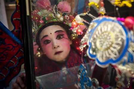 A child in red traditional costume with bright pink face makeup and decorated headdress gazes at her reflection in a mirror surrounded by elaborate props.