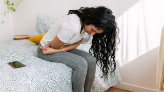 A young woman sits crouched over on a bed with her arms hugging her lower belly like she is in a lot of pain