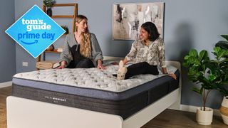 Two women sat on Helix Midnight Luxe mattress in sleep studio with green plants and shelving either side of bed and Tom's Guide Prime Day