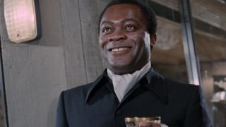 Yaphet Kotto stands smiling, with a drink in his hand, in Live and Let Die.