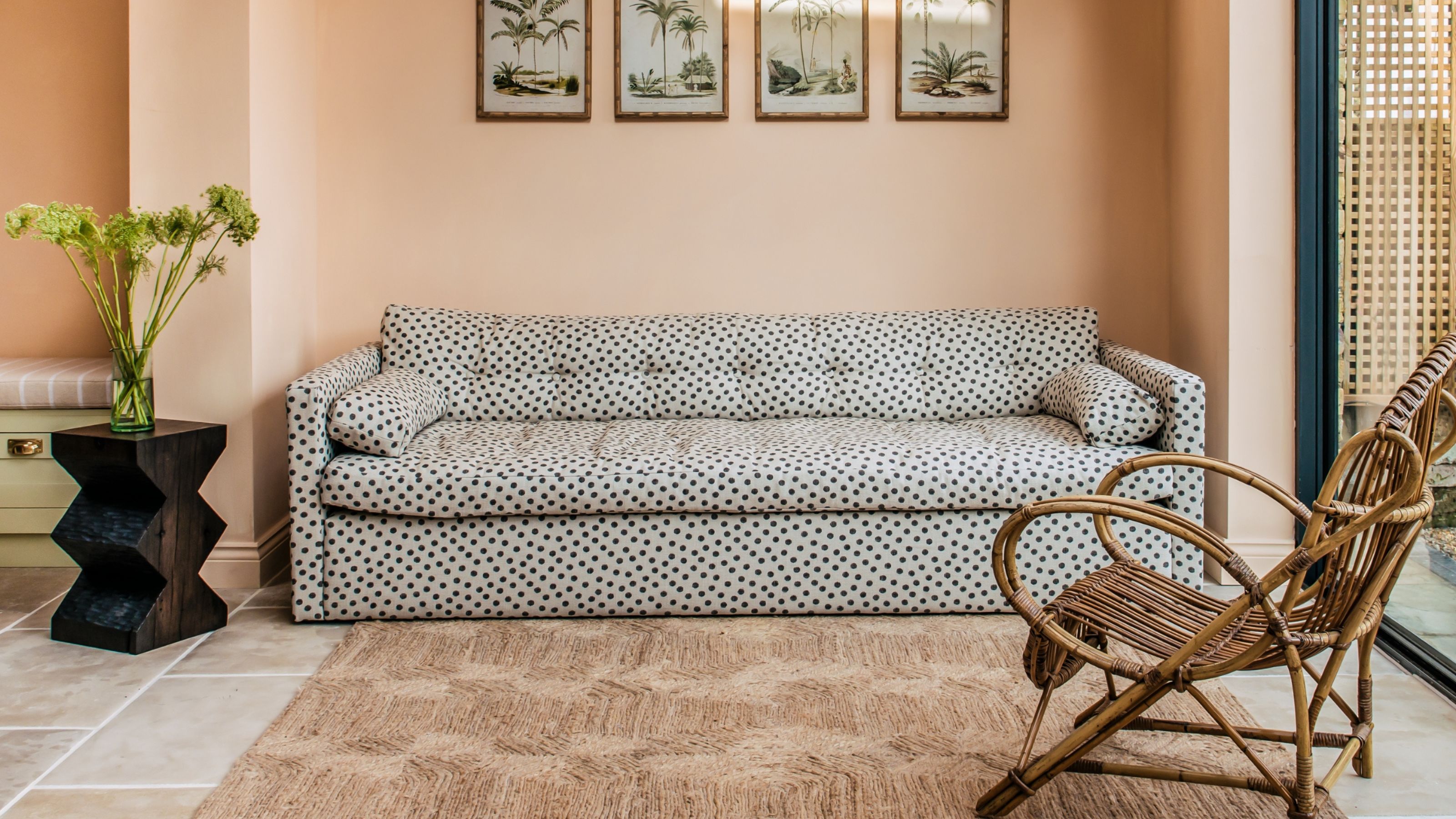 living room that is painted peach and has large tile floors. There is a black and white polka dot sofa and a wicker armchair. There are four paintings on the wall above the sofa and a jute rug on the floor.