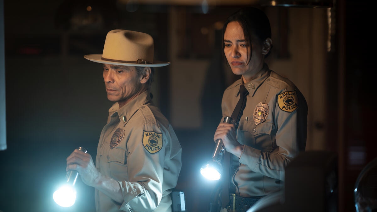 Zahn McClarnon as Lieutenant Joe Leaphorn and Jessica Matten as Sergeant Bernadette Manuelito wearing their police uniform and shining torches at something on the floor.