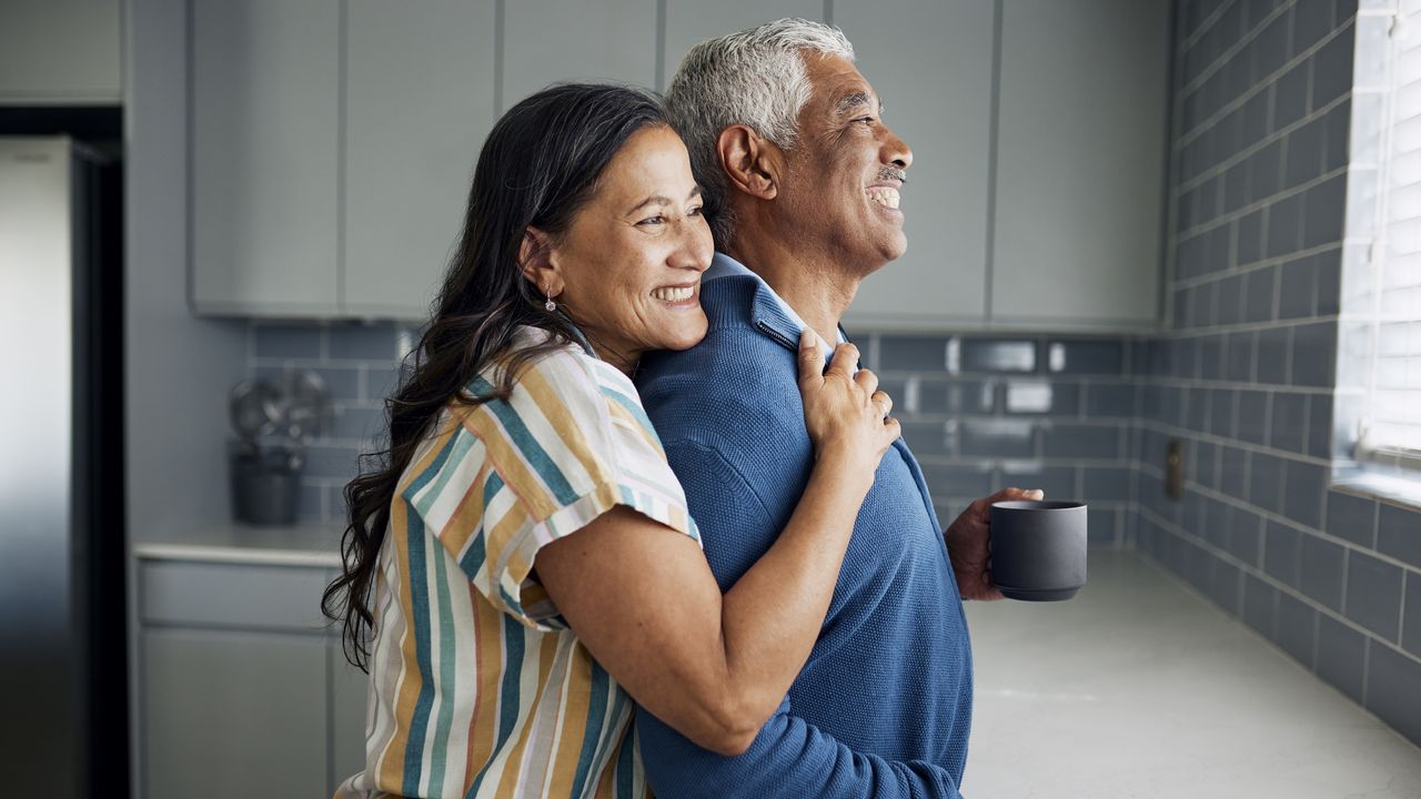 A happy retired couple hug in the kitchen while looking out the window.