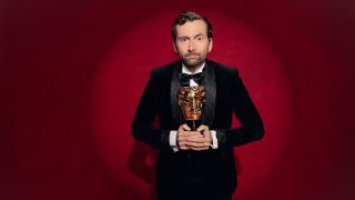 David Tennant in a suit holding a golden BAFTA trophy and standing in a spotlight in front of a red curtain