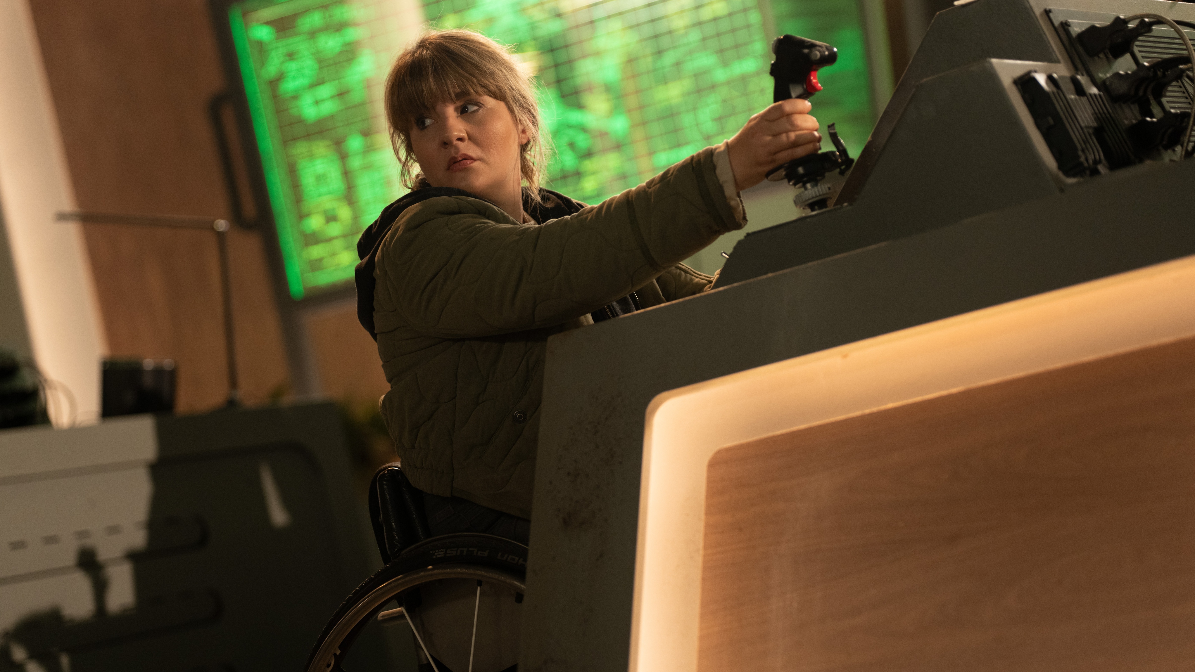 Shirley Bingham (Ruth Madeley) is at her desk in UNIT HQ, in her wheelchair, with her right hand holding a joystick-style controller. There is a screen behind her showing some sort of map information in green. She is wearing an anorak over a hooded top.