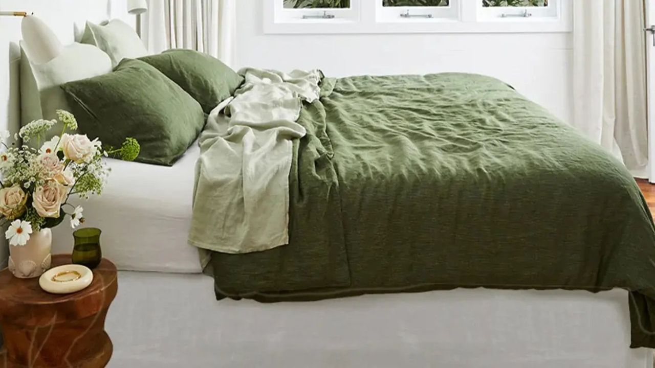 Side view of olive and sage sheets on a bed against white walls; in the foreground, a dark wood nightstand topped with a vase of flowers. 