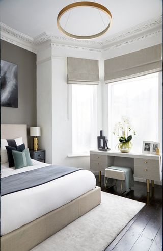 neutral bedroom with gold circular pendant light, dressing table, bay window blinds and dark wood flooring