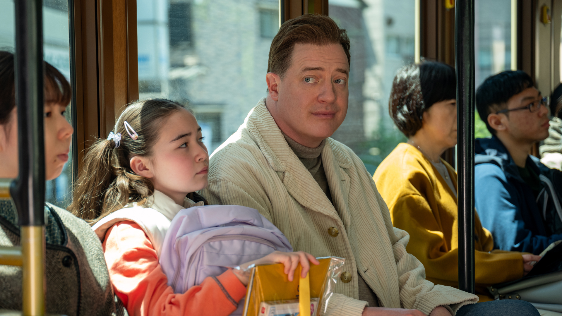 A group of people sitting on a bus. A young girl (Shannon Gorman as Mia) holds a backpack and yellow school supplies, while a man (Brendan Fraser as Phillip) sits next to her, wearing a tan coat. A film still from 'Rental Family.'
