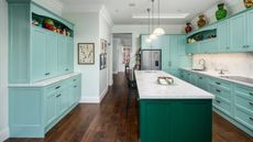 Teal and green kitchen with kitchen island, wooden floors, three hanging lights. Vases and ornaments are positioned on the shelves and above cabinets, and a hallway is pictured in the background.