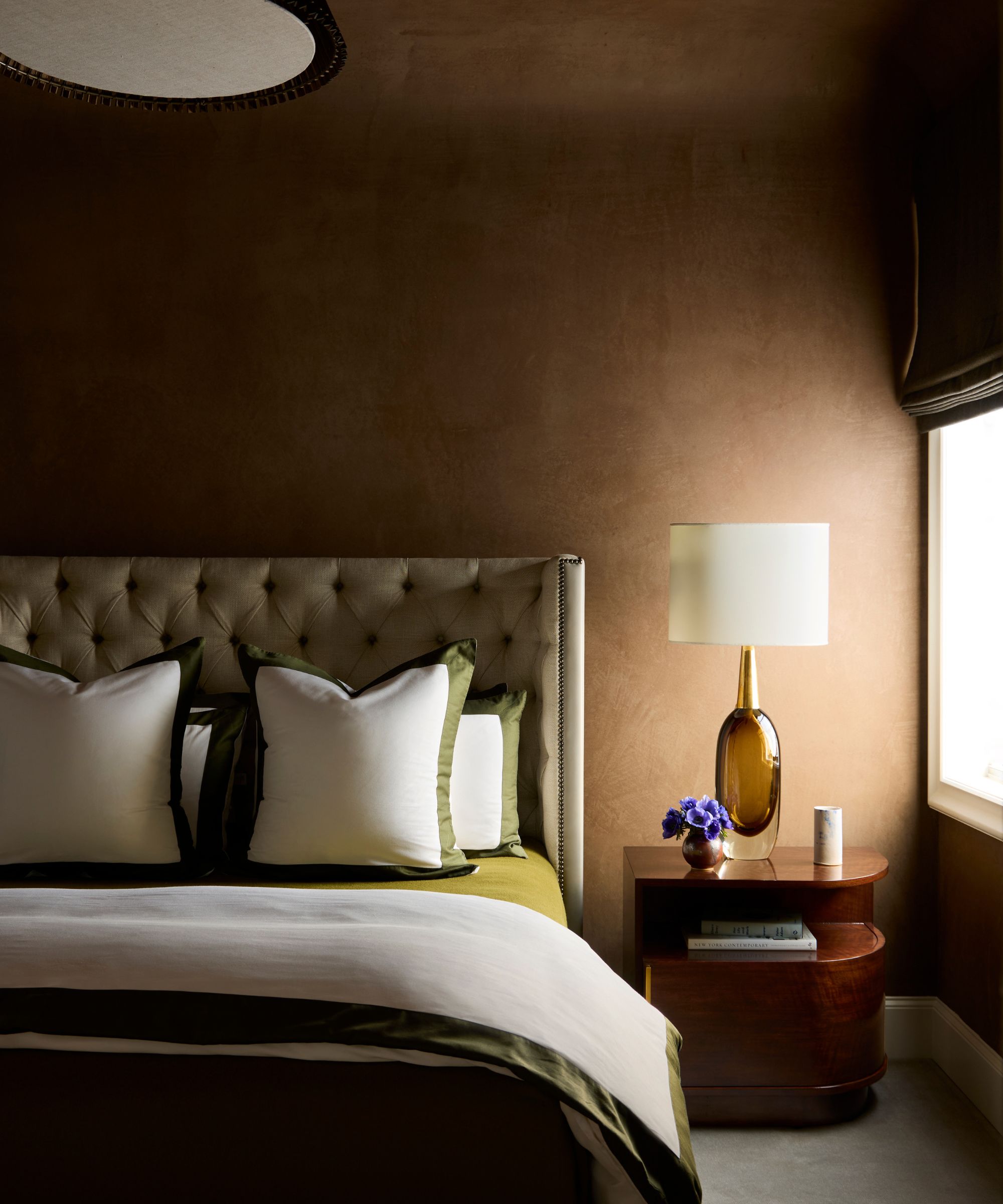 A bedroom with brown color-drenched walls in a plaster finish. A bed with a gray headboard and white bedding with green trim.