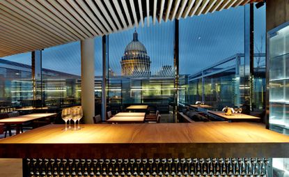 A restaurant with a large wooden counter with glasses underneath it, rows of dining tables, glass walls and a view of the dome of St Isaac’s Cathedral.