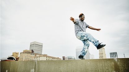 A young man walks along the edge of a roof looking like his balance is precarious.