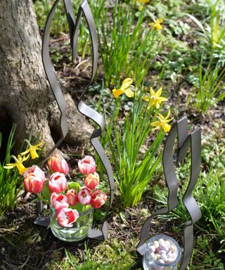 Two metal spring bunny decorations - one with a glass jar of tulips and one with a glass jar of pastel eggs, on green grass with daffodils, next to a tree