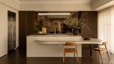 A white kitchen with brown wood cabinets, a textured backsplash, a white dual deck island, wood chairs, a black vase with stems, a bowl of fruit, and a leaning painting