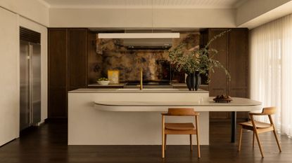 A white kitchen with brown wood cabinets, a textured backsplash, a white dual deck island, wood chairs, a black vase with stems, a bowl of fruit, and a leaning painting