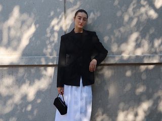 dominique wears black blazer, white skirt, black mesh flats, and black handbag while leaning against a wall