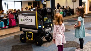 Two small children stood looking at one of Ottonomy's Ottobots, autonomous delivery vehicles created as an xBridge pilot project, within a terminal at Pittsburgh International Airport. The Ottobot is a large box with a window that can be opened to fit packages, with two eye-shaped lights on the front and four wheels.