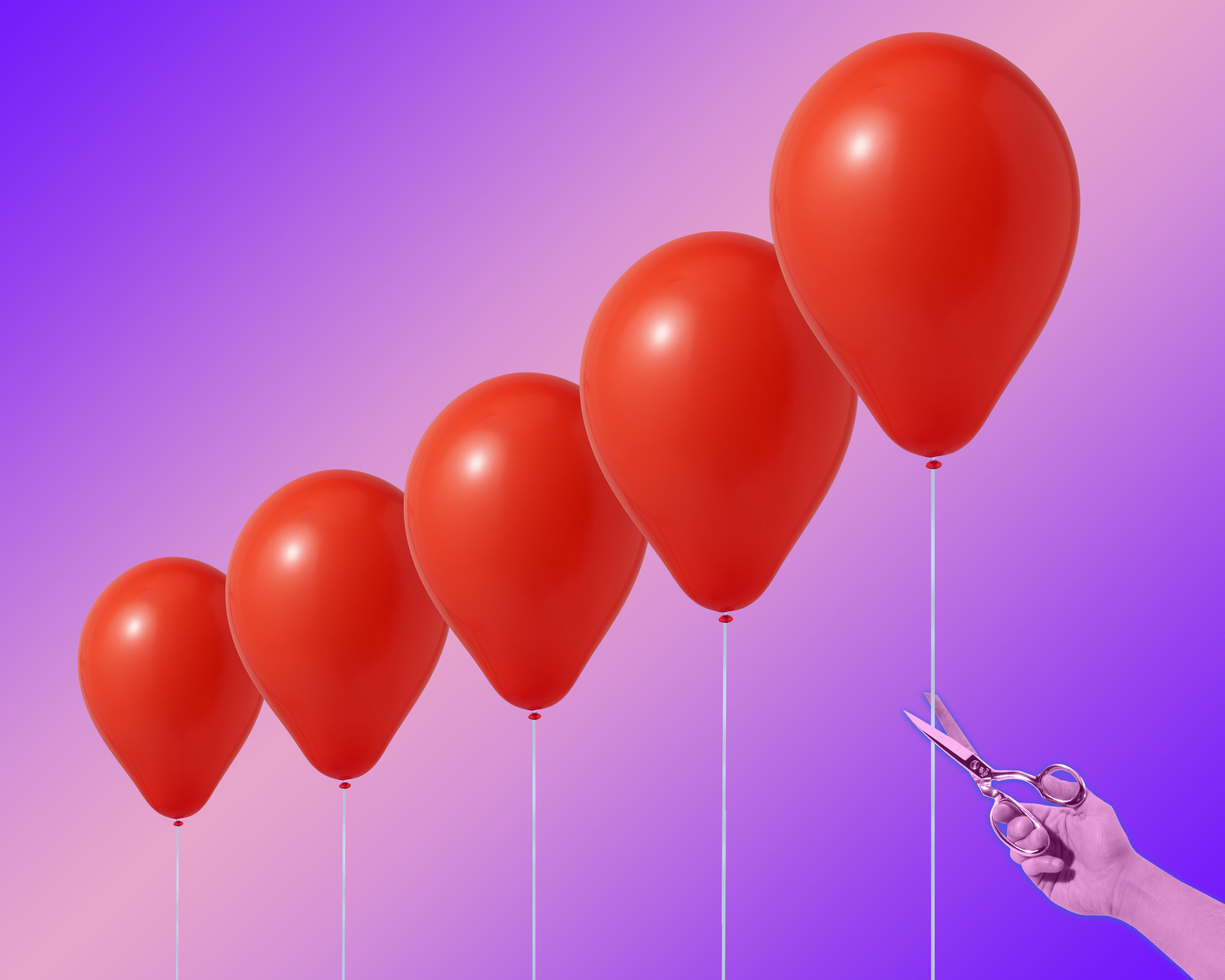 Man's hand with steel scissors about to cut the string of one of 5 variously sized red helium balloons, with a gradated purple background