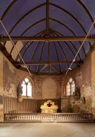 Interior view of Château de Boisgeloup featuring distressed walls, wood beams, an arched roof, wall mounted statues and two window frames, one of which has stained glass. There is a light coloured decorative table on the floor with a red cross and other items on top