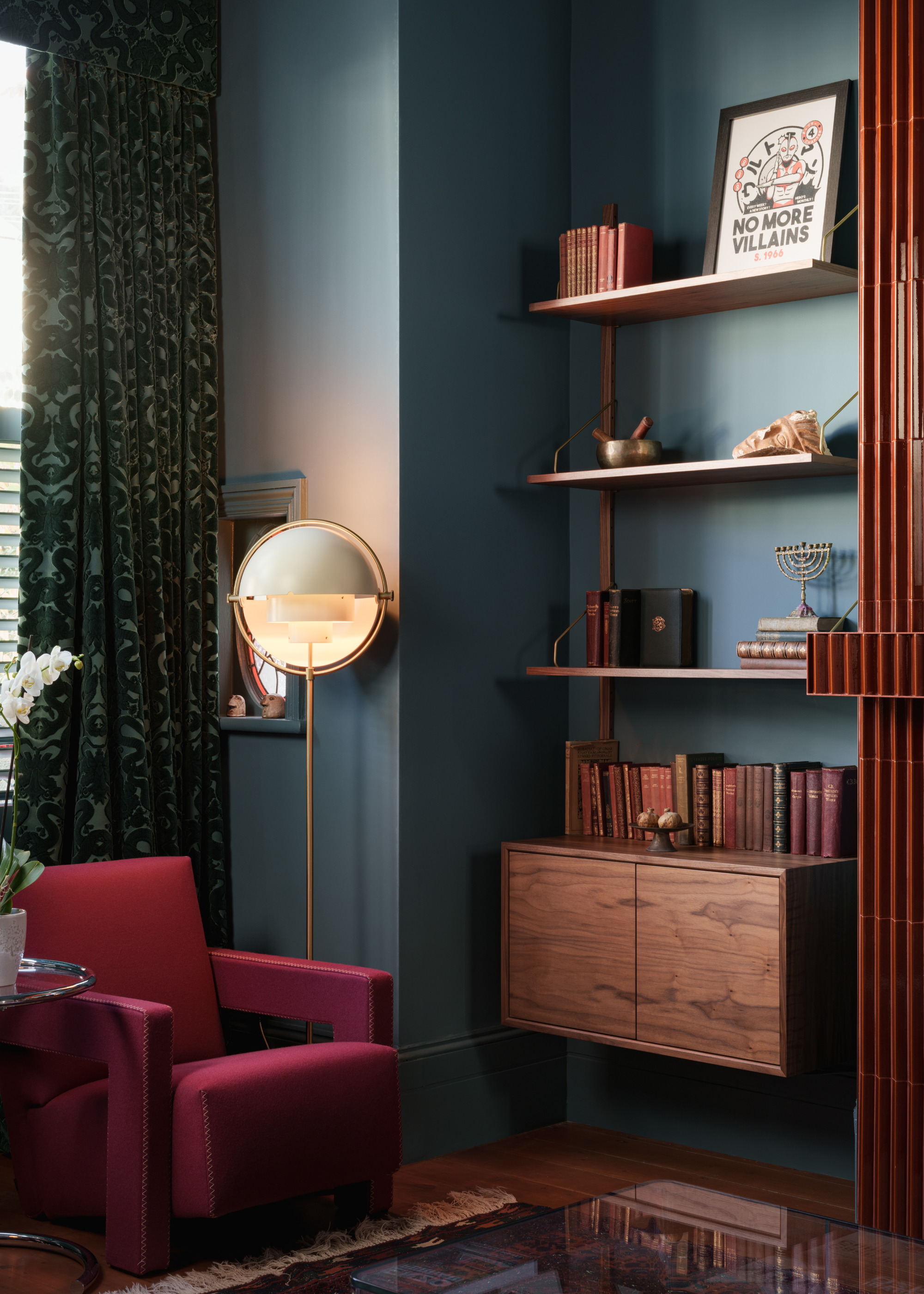 A living room corner with a red accent chair in front of a window with floor-length patterned dark green curtains, a round orb floor lamp and open shelving filled with books and decorative objects