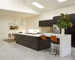 Open plan kitchen with neutral, natural color palette, gray tiled flooring, large central kitchen island and breakfast bar, black cabinets and white countertops, brown leather bar stools with black metal frames, separate dining area to the left, textured pendants hanging over seating area