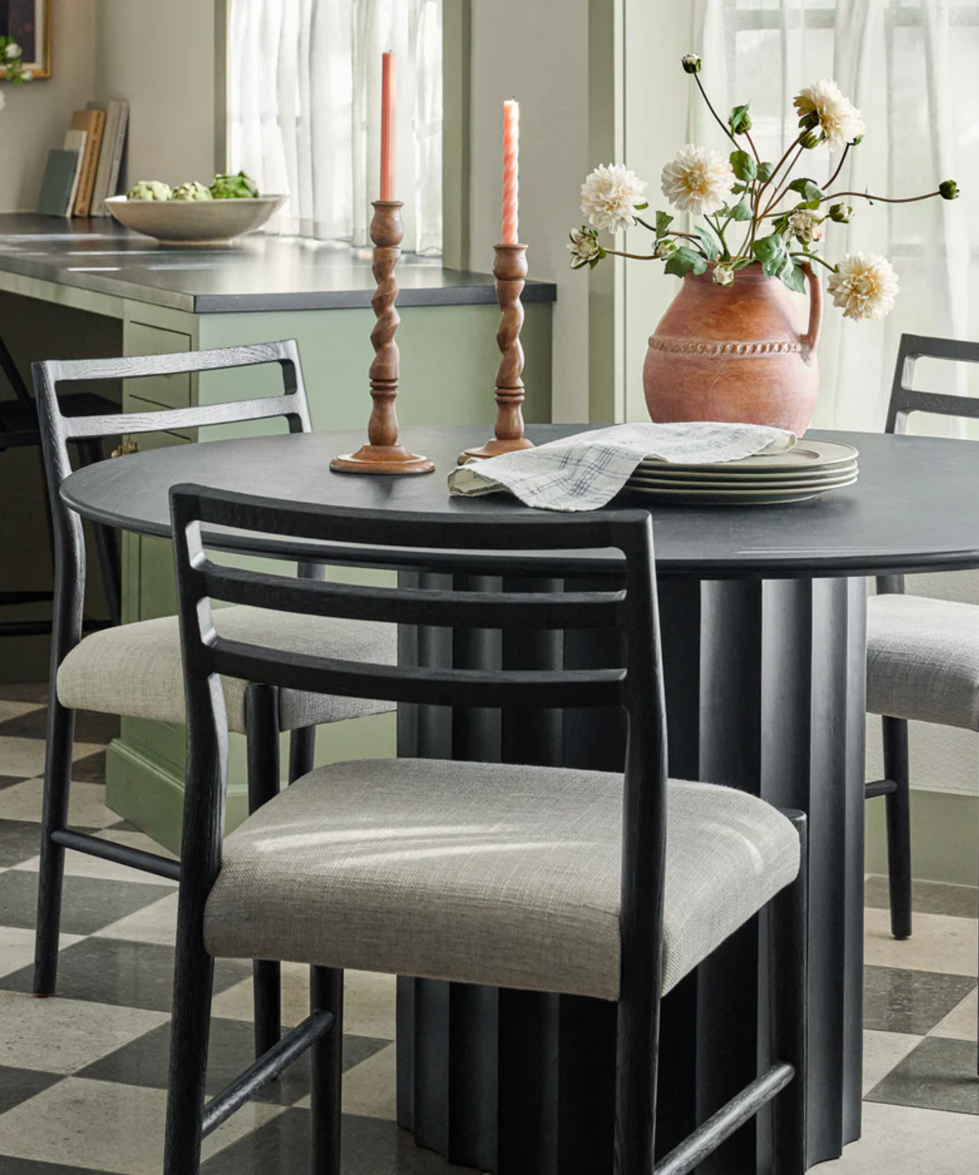 a dining room with a black dining table and dining chairs, twisted wood candle holders, a set of plates, and a vase holding flowers