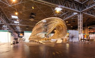 The Twist at the Timber Expo 2015 by the Architectural Association, exhibition hall, wooden floor, metal strut high ceiling with skylights and lighting, wooden sculpture in the centre, visitor guide rope to the left and right, metal pillar, blurred people images walking by